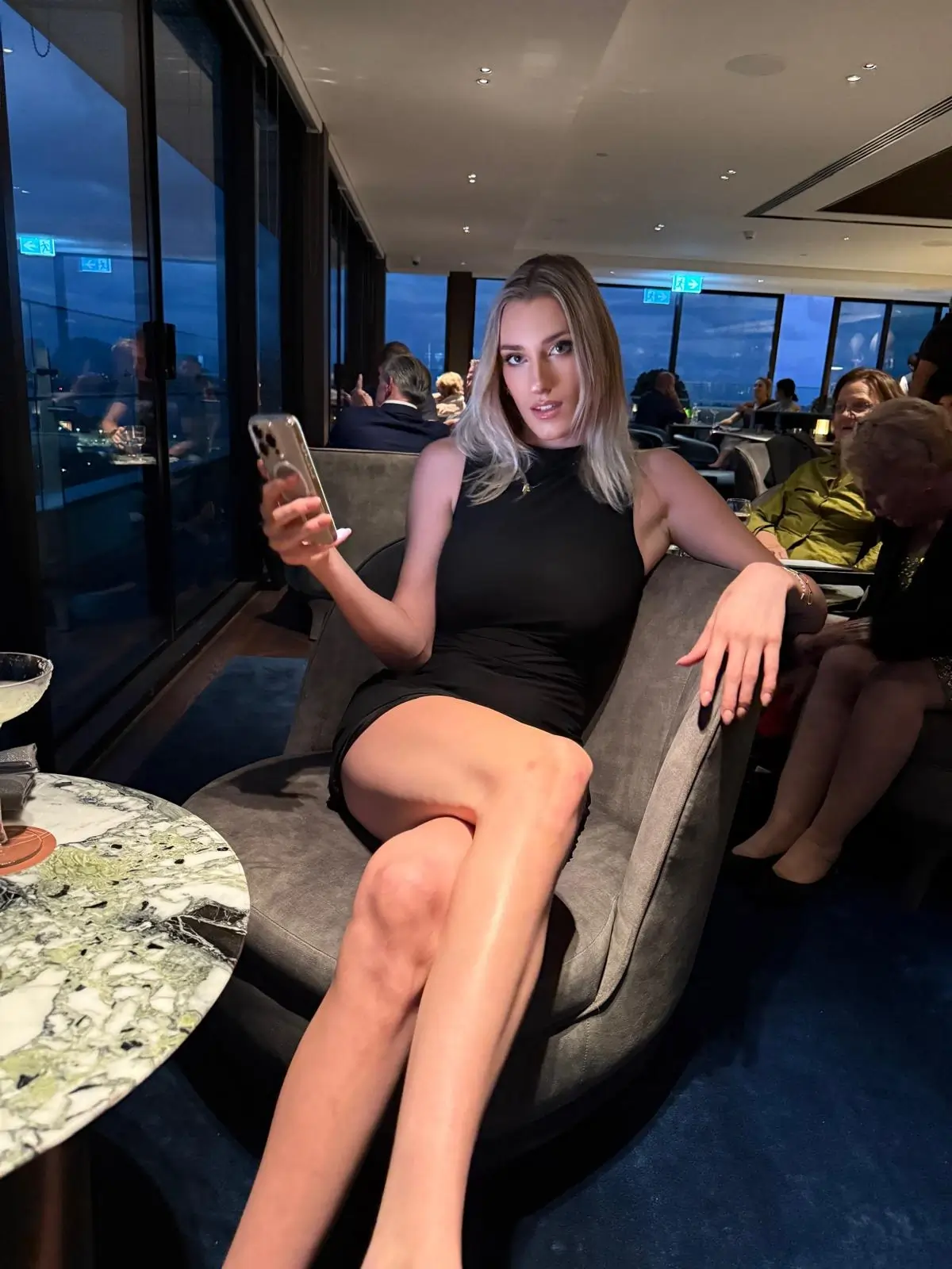 Serena in a sleek black dress relaxing in a lounge bar, holding her phone with city views behind.