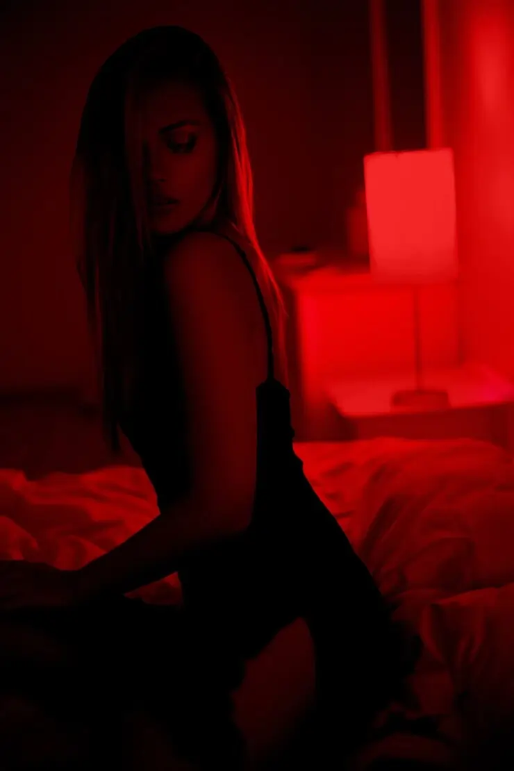 Sensual woman in a dark bedroom illuminated by red lighting, sitting on a bed with soft shadows creating a moody, intimate atmosphere.
