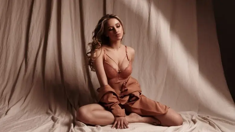 Woman sitting on fabric in soft natural light, wearing matching lingerie and an open silk robe, with a calm and intimate expression.
