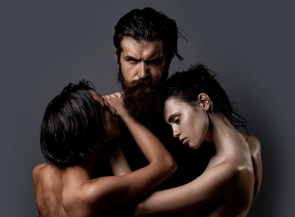 Bearded man embracing two topless women on a dark studio backdrop, intimate trio composition.
