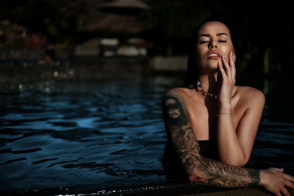 tattooed brunette in a dark swimsuit emerging from a pool at night with one hand on her face