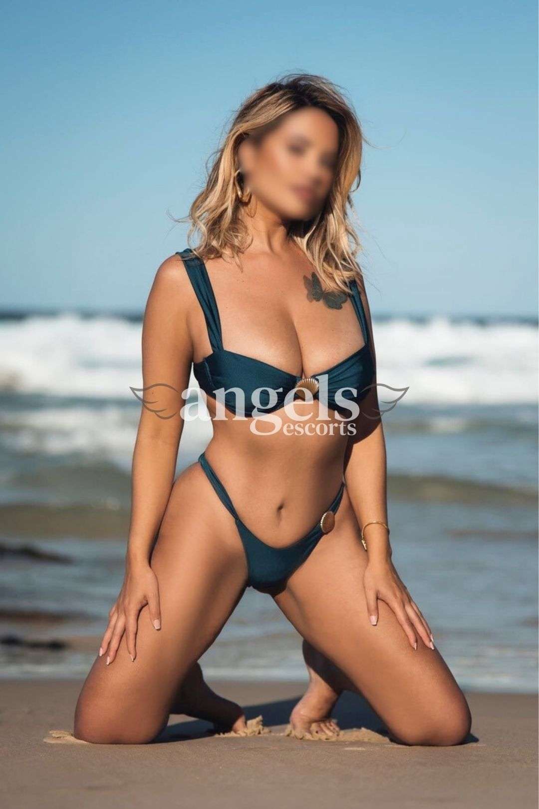 Irina kneeling on the beach in a deep green bikini with waves and blue sky behind her.